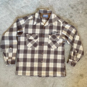 May include: A brown and white plaid flannel shirt with a long sleeve and a button-up front. The shirt has a patch pocket on the left chest and a small embroidered logo that says "The Bakery" on the right chest.