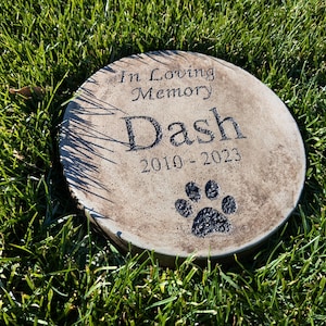 May include: A round, gray concrete memorial stone with the inscription "In Loving Memory Dash 2010-2023" and a black paw print.