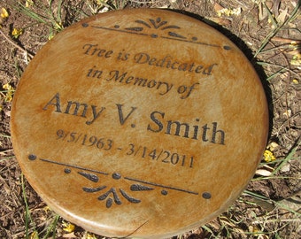 May include: A brown, round, engraved memorial stone with the text "Tree is Dedicated in Memory of Amy V. Smith 9/5/1963-3/14/2011".