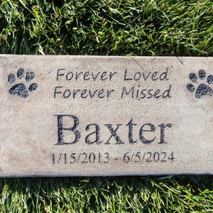 May include: A light brown rectangular stone memorial with black paw prints and the text "Forever Loved Forever Missed" and "Baxter 1/15/2013 - 6/5/2024".