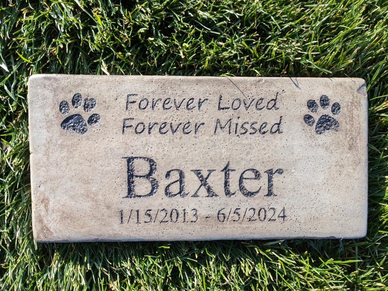May include: A light brown rectangular stone memorial with black paw prints and the text "Forever Loved Forever Missed" and "Baxter 1/15/2013 - 6/5/2024".