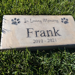 May include: A light brown rectangular concrete memorial stone with the text "In Loving Memory Frank 2010-2021" and two paw prints.
