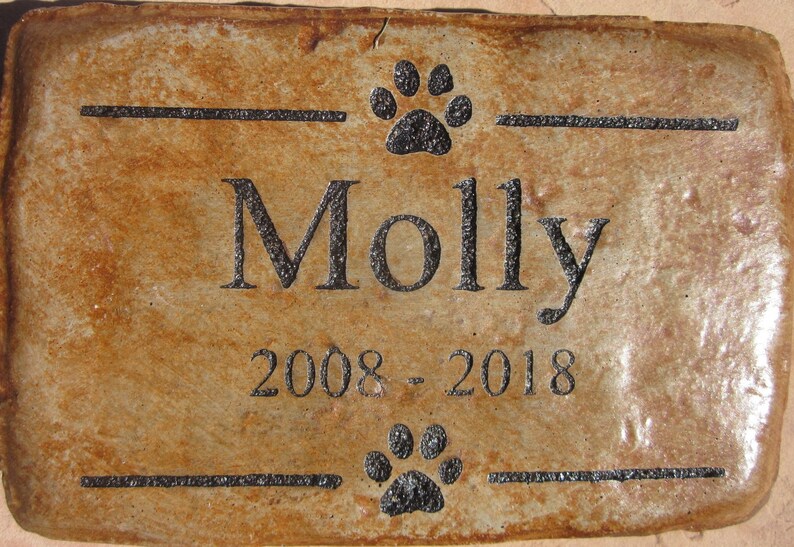 May include: A brown rectangular stone memorial with black engraved text. The text reads "Molly 2008 - 2018" with a paw print above and below the name.