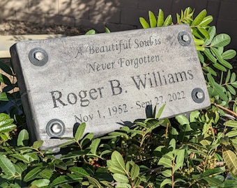 May include: A gray concrete memorial plaque with rounded corners and a black inscription that reads "A Beautiful Soul is Never Forgotten Roger B. Williams Nov 1, 1952 - Sept 27, 2022". The plaque has three silver metal fasteners.