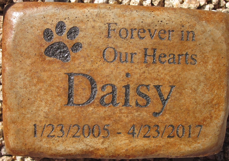 May include: A brown rectangular stone memorial with a black paw print, the text "Forever in Our Hearts", the name "Daisy", and the dates "1/23/2005 - 4/23/2017".