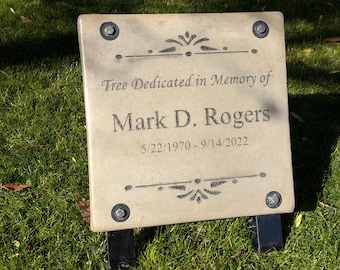 May include: A beige concrete plaque with rounded corners and four silver screws. The plaque is mounted on a black metal stand. The plaque reads "Tree Dedicated in Memory of Mark D. Rogers 5/22/1970 - 9/14/2022".