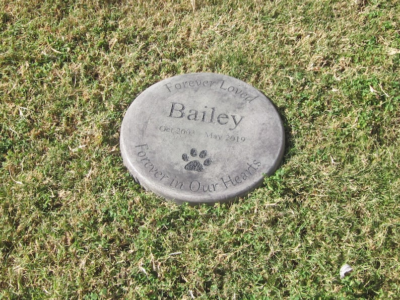 以下が含まれることがあります： 「Forever Loved Bailey Oct 2003 - May 2019 Forever in Our Hearts」と肉球の跡が刻まれた灰色のコンクリート製の記念碑