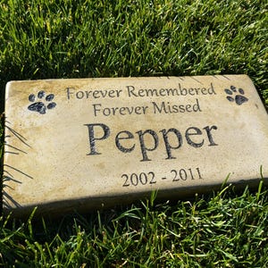 May include: A beige concrete memorial stone with black paw prints and the inscription "Forever Remembered Forever Missed Pepper 2002-2011".