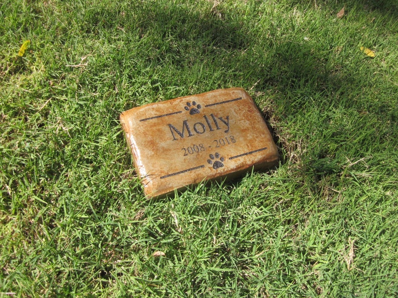 May include: A brown rectangular stone memorial with the name "Molly" and the dates "2008 - 2018" engraved on it.  The stone is surrounded by green grass.