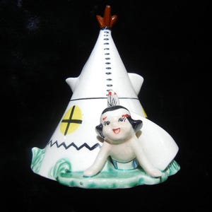 May include: A vintage ceramic figurine featuring a white teepee with a yellow cross-shaped window and a smiling Native American figure. The figurine has a green base with white waves and a brown top.