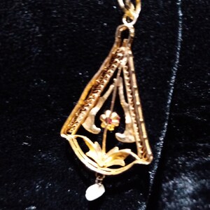 May include: A gold pendant featuring a delicate floral design with a single red gemstone. The pendant hangs from a thin gold chain and has a small pearl drop.