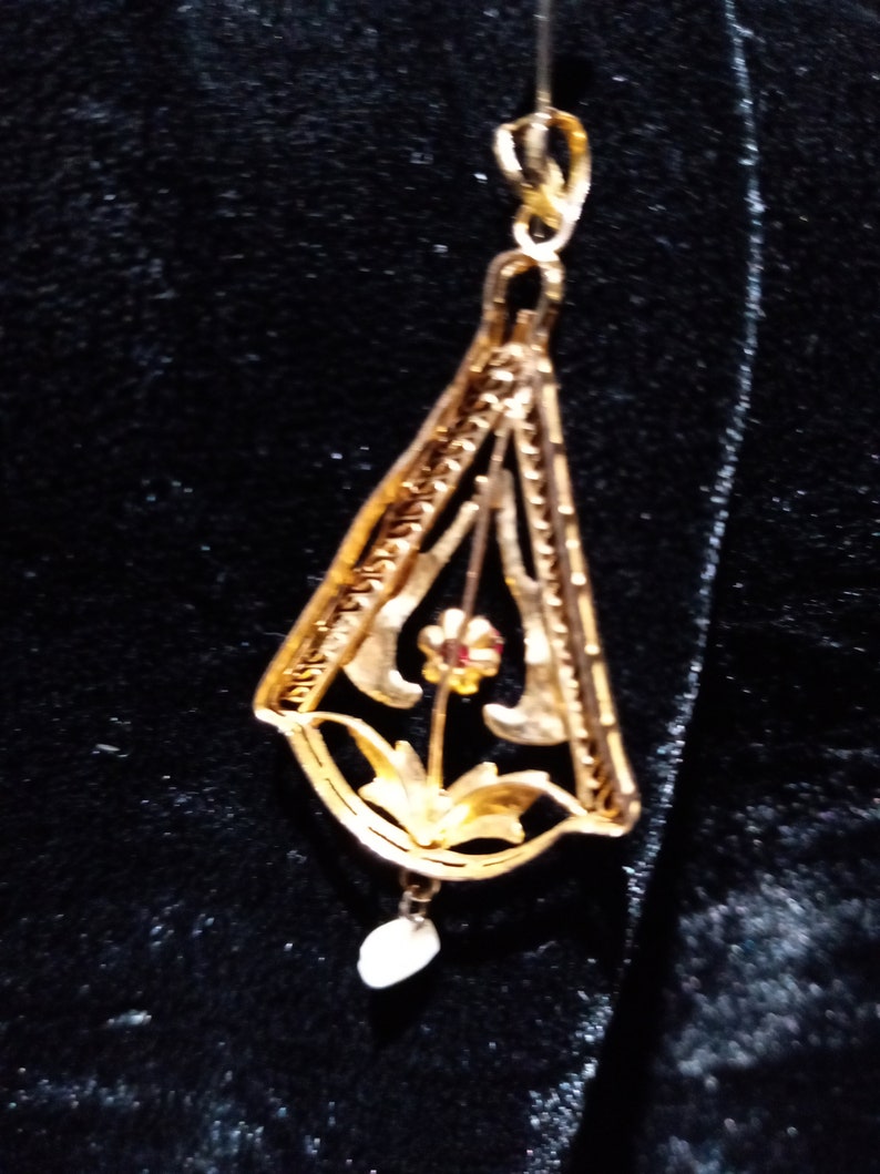 May include: A gold pendant featuring a delicate floral design with a single red gemstone. The pendant hangs from a thin gold chain and has a small pearl drop.
