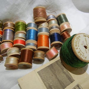May include: A collection of spools of thread in various colors, including green, blue, red, and brown. The spools are arranged in a semi-circle on a white surface. A spool of green thread with the text "PURE EXTRA QUALITY ENGLISH SILK MOTOR ALICE STEWART NEW YORK" is in the foreground.