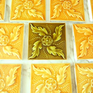 May include: A collection of square ceramic tiles featuring a raised relief pattern of a central flower surrounded by swirling acanthus leaves. Most tiles are a bright yellow-orange, while one central tile is a deep olive green.