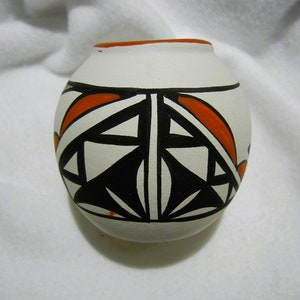 May include: A small, white ceramic pot with a geometric design in black and orange. The pot is round and has a slightly flared lip.