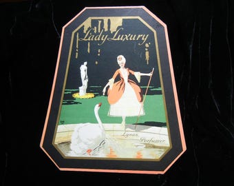 May include: Vintage Lady Luxury perfume advertisement. The artwork features a woman in a period dress, a swan, and a statue in a garden setting. The frame is black with gold accents and a peach border. The text "Lady Luxury" is at the top, and "Lynas, Perfumer" is at the bottom.