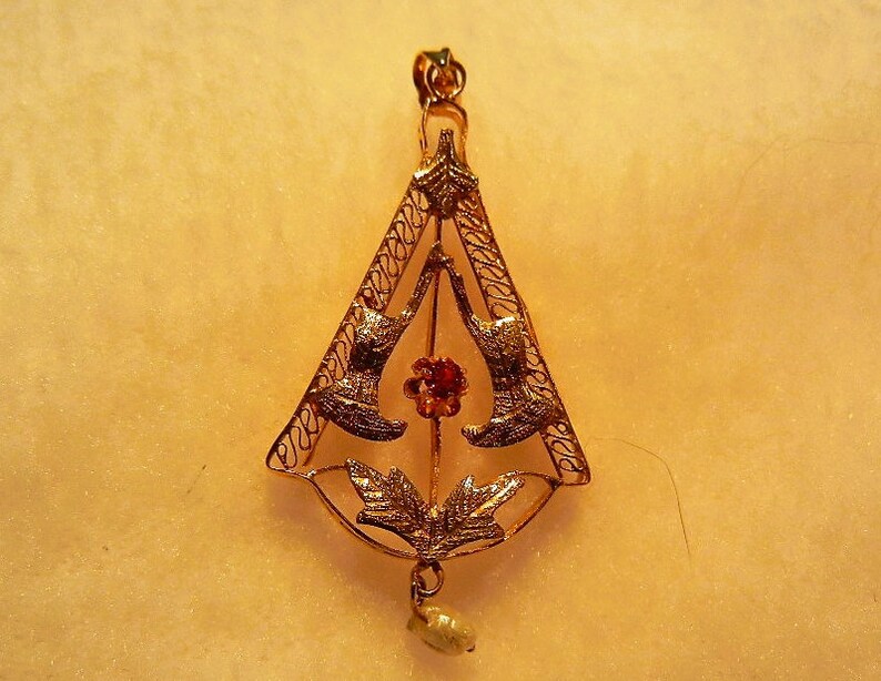 May include: A gold pendant with a delicate filigree design featuring two birds facing each other. The pendant has a red gemstone in the centre and a pearl drop at the bottom.