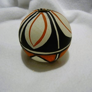 May include: A small, round, white ceramic pot with black and orange geometric designs. The pot has a small opening at the top.