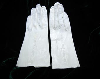 May include: A pair of white leather gloves. The gloves have a classic design with decorative stitching and small cut-out details on the back of the hands. The gloves are displayed against a dark background.