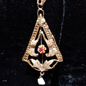 May include: A gold pendant with a delicate filigree design featuring two birds and a red gemstone. The pendant is suspended from a gold chain and has a small pearl drop.