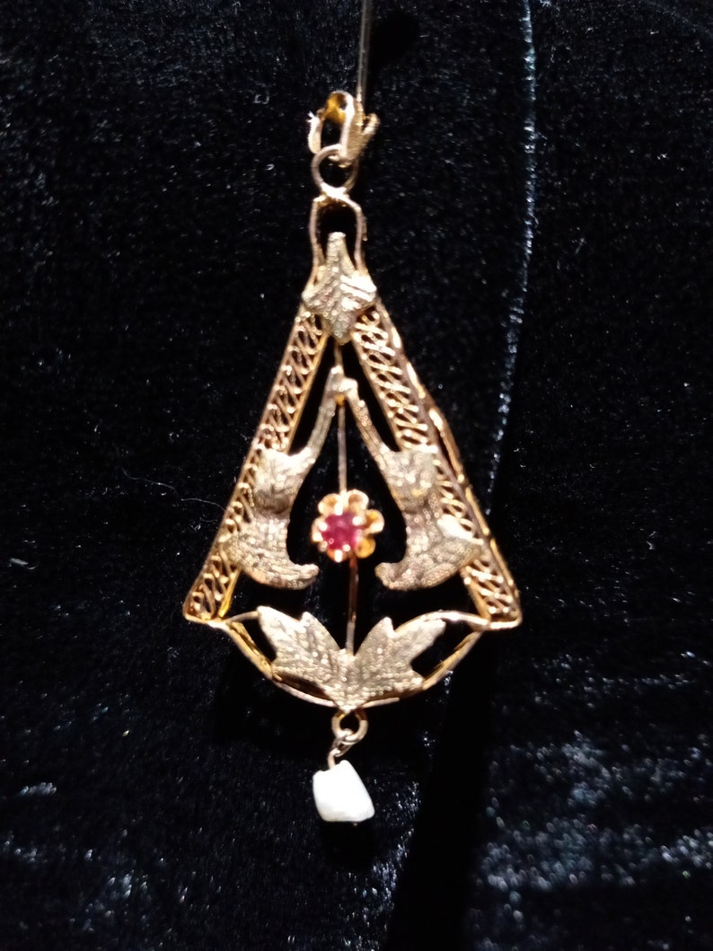 May include: A gold pendant with a delicate filigree design featuring two birds and a red gemstone. The pendant is suspended from a gold chain and has a small pearl drop.