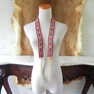 May include: A long, flat beaded neckpiece featuring a geometric pattern in red, black, and white. The beaded section is attached to two light-colored leather fringe strips that hang down from the bottom of the beaded band on each side.