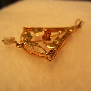 May include: A gold pendant with a delicate design featuring leaves and a red gemstone. The pendant is attached to a chain with a pearl-like bead.