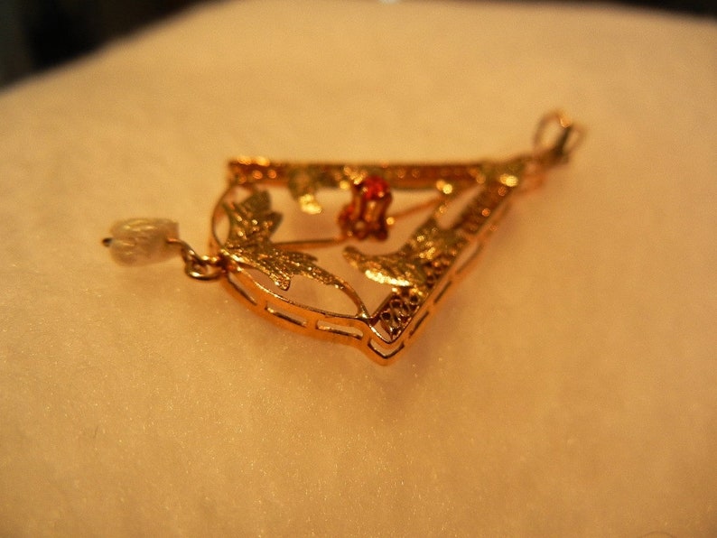 May include: A gold pendant with a delicate design featuring leaves and a red gemstone. The pendant is attached to a chain with a pearl-like bead.