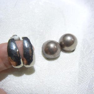 May include: A pair of silver hoop earrings and two silver ball stud earrings. The hoop earrings are on a finger, and have a hammered texture. The stud earrings are smooth and round. The jewelry is displayed on a white, textured surface.