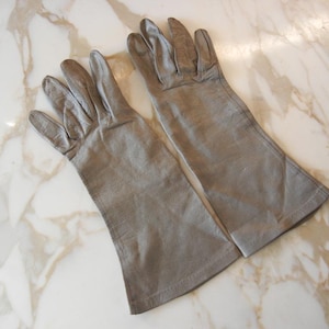 May include: A pair of light gray leather gloves, laid flat on a marble surface. The gloves have a classic design with long sleeves and are made of a soft, supple material. The gloves are a neutral color, suitable for various outfits.