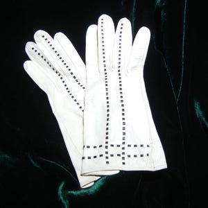 May include: A pair of white gloves with black square stitch detailing. The gloves are made of a smooth material and have a classic design. The stitching is visible on the fingers and the back of the gloves. The gloves are likely for formal wear.