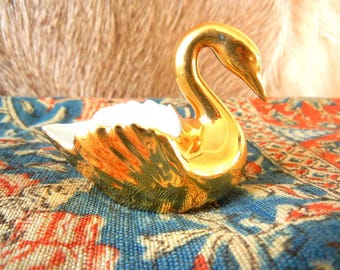 May include: A gold-colored ceramic swan figurine with a curved neck and detailed wing textures. The interior of the swan is white, and the exterior features a reflective, metallic gold finish that catches the light, highlighting the sculpted feathers.