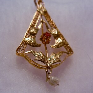 May include: A gold pendant featuring a delicate design of leaves and a red gemstone. A single pearl hangs from the bottom of the pendant.
