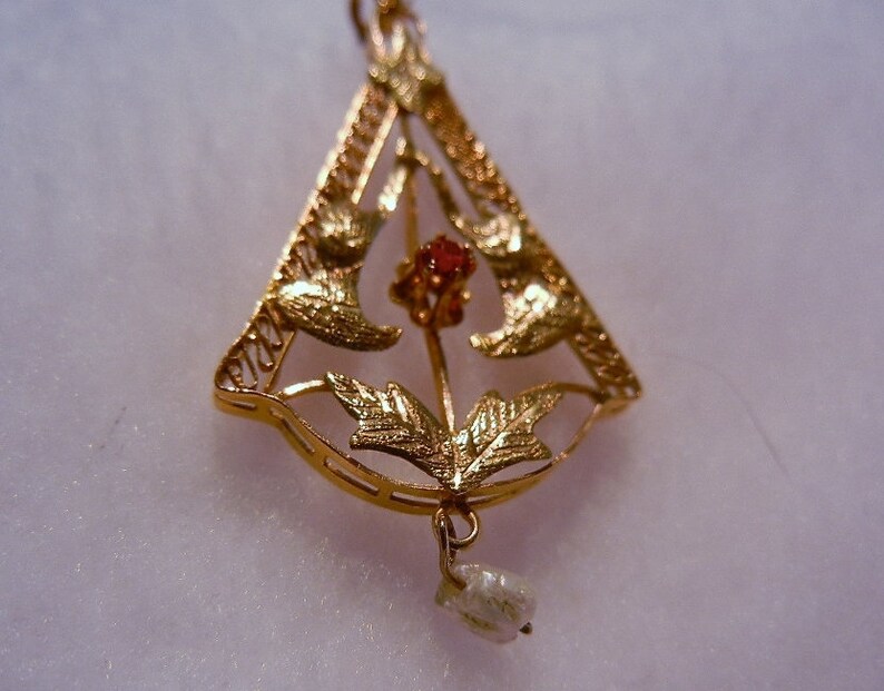 May include: A gold pendant featuring a delicate design of leaves and a red gemstone. A single pearl hangs from the bottom of the pendant.