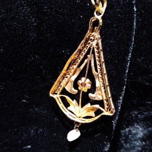 May include: A gold pendant with a delicate filigree design featuring a flower with a red centre. The pendant hangs from a thin gold chain and has a small pearl drop.