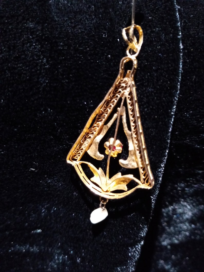 May include: A gold pendant with a delicate filigree design featuring a flower with a red centre. The pendant hangs from a thin gold chain and has a small pearl drop.