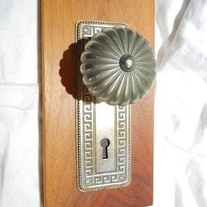 May include: Antique door hardware featuring a silver-toned, fluted doorknob and a rectangular backplate with a keyhole. The backplate has a decorative Greek key border and is mounted on a rectangular wooden board.