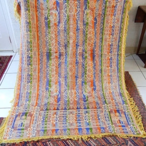 May include: A rectangular woven throw blanket featuring vertical stripes in orange, blue, and green. The fabric displays intricate floral patterns, architectural motifs, and animal figures throughout. The edges are finished with yellow fringe.