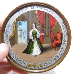May include: A round, antique miniature painting with a gold-toned frame. The painting depicts a woman in a green and white dress in a room with red curtains and a red chair. The text "Mlle de la Valliere" is at the top.