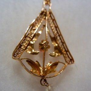 May include: A gold pendant with a delicate filigree design featuring a floral motif. The pendant is shaped like a triangle and has a small red stone in the centre.