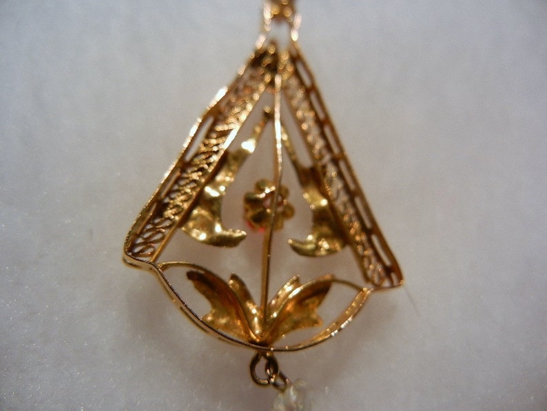 May include: A gold pendant with a delicate filigree design featuring a floral motif. The pendant is shaped like a triangle and has a small red stone in the centre.