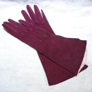 May include: A pair of deep burgundy gloves with a smooth, matte finish. The gloves feature stitched detailing along the back of the hand and fingers, with a flared cuff design that reveals a light-colored interior lining at the wrist opening.