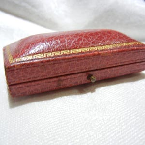 May include: A small, rectangular, red jewelry box with a textured surface. The box has a gold-colored decorative border and a small, round clasp. The box is closed and sits on a white surface.