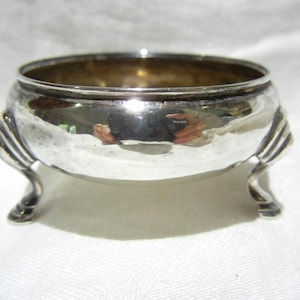 May include: A small, antique silver bowl with a rounded body and three curved feet. The bowl has a wide, open top and a reflective surface. The feet are decorative, with a ribbed design. The bowl is likely used for serving or display.