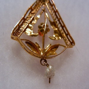 May include: A gold pendant featuring a delicate floral design and a single pearl drop. The pendant is crafted from filigree and has a triangular shape.