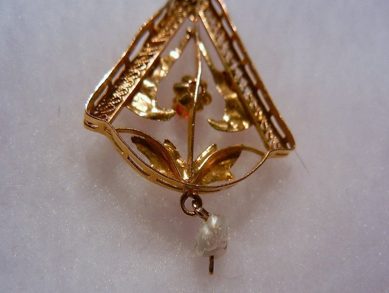 May include: A gold pendant featuring a delicate floral design and a single pearl drop. The pendant is crafted from filigree and has a triangular shape.