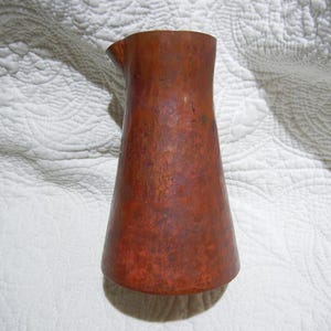 May include: A copper-colored pitcher with a flared base and a narrow spout. The pitcher has a textured surface and a warm, reddish-brown hue. The design is simple and elegant, suitable for serving beverages.
