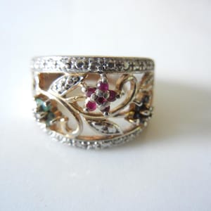 May include: A wide band ring featuring a gold-tone metal finish with openwork floral motifs. The design includes small embedded red, green, and dark blue gemstones arranged in flower shapes, accented by tiny clear stones along the top and bottom edges.