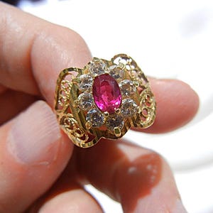 May include: A gold ring featuring a central oval-cut vibrant pink gemstone surrounded by a cluster of eight clear round-cut stones. The band displays an intricate openwork filigree design with textured gold detailing on the sides.