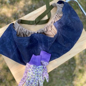 May include: A navy blue gardening bag with a green strap and a pair of gardening gloves. The gloves have purple cuffs and a floral pattern with green and purple flowers. The bag is made of a waxed canvas material.